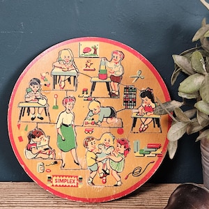 Op de afbeelding: Een ronde houten puzzel met een kleurrijke illustratie van kinderen in een klaslokaal met een leraar. De puzzelstukjes tonen diverse activiteiten zoals spelen met blokken, lezen en tekenen. Het woord SIMPLEX staat onderaan afgedrukt.