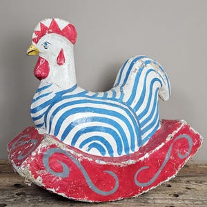 Op de afbeelding: Een wit haanfiguur met blauwe strepen op het lichaam, een rode kam en een gele snavel. De haan is gemonteerd op een gebogen rode schommelbasis versierd met grijze wervelpatronen. Het oppervlak van het object vertoont tekenen van slijtage.
