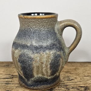 Op de afbeelding: Een handgemaakte steengoed mok met een getextureerd oppervlak. De mok heeft een donkerblauw en bruin kleurenschema, een afgeronde body en een stevige handgreep. De binnenkant is donkerbruin.