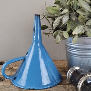 Op de afbeelding: Een felblauwe trechter van geëmailleerd metaal met een brede conische vorm, een smalle tuit en een gebogen handvat aan de zijkant. Het oppervlak vertoont lichte gebruikssporen met kleine donkere vlekjes bij de tuit en de rand.
