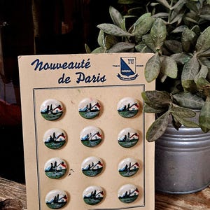 Op de afbeelding: Een set van twaalf ronde knopen op een beige displaykaart met het opschrift Nouveauté de Paris. Elke knoop is voorzien van een handgeschilderd landschap met een klein huisje, groen gras en een blauwe lucht. De kaart bevat een logo en de tekst Fabrication Française.