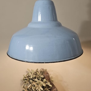 Op de afbeelding: Een lichtblauwe hanglamp in vintage-stijl met een donkere metalen fitting. De lamp hangt boven een rustiek houten oppervlak, met een kleine metalen emmer met groen en een bezem. Er is ook een linnen doek met Franse tekst aanwezig.