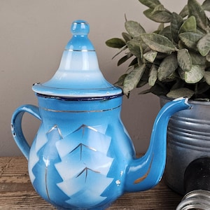 Op de afbeelding: Een vintage blauw-witte geëmailleerde theepot met een puntig deksel met zilveren rand en een gebogen tuit. Het lichaam heeft een herhalend wit geometrisch patroon met zilveren accenten. De emaille afwerking vertoont lichte gebruikssporen.