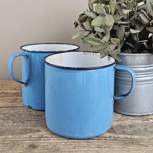Puede incluir: Dos tazas de esmalte azul claro con interiores blancos y bordes negros finos. Cada taza cuenta con un asa curva unida al lateral. El acabado exterior es liso y de color azul sólido, mientras que el interior muestra una superficie blanca.