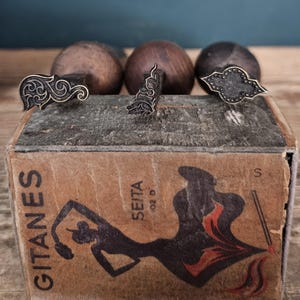 Puede incluir: Tres herramientas de estampado de metal con mangos de madera oscura están colocadas sobre una caja de cartón rectangular. La caja presenta el texto GITANES y SEITA, junto con una ilustración gráfica negra de una persona bailando con detalles rojos.