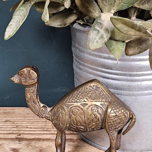 May include: A brass camel figurine featuring intricate etched patterns across its body, including geometric cross-hatching and circular panels depicting seated figures. The metal surface has a dark, aged patina with visible highlights.