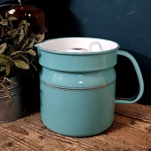 Op de afbeelding: Een cilindrische turquoise geëmailleerde kan met een witte binnenkant, een enkel handvat aan de zijkant en een dunne zilverkleurige horizontale band rond het bovenste gedeelte. De kan heeft een wit deksel met een metalen lusgreep.