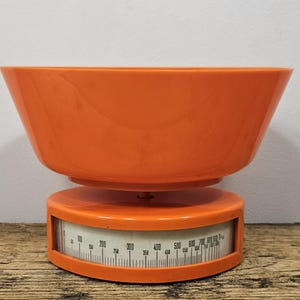 Op de afbeelding: Een feloranje keukenweegschaal met een grote, diepe, bijpassende oranje kom op een ronde voet. De voet bevat een gebogen, wit displayvenster met zwarte numerieke markeringen variërend van 0 tot 1 kilogram voor nauwkeurig wegen.