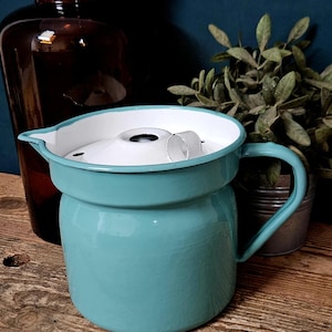 Op de afbeelding: Een turquoise geëmailleerde kan met een witte binnenkant en een bijpassend wit deksel. De kan is voorzien van een gebogen handvat aan de zijkant, een kleine schenktuit en een ronde greep bovenop het deksel om het gemakkelijk te verwijderen.