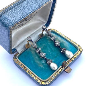 Può includere: Un paio di orecchini pendenti antichi esposti in un cofanetto blu. Gli orecchini presentano un design in metallo scuro con pietre preziose trasparenti e perle a goccia. La scatola ha un interno in velluto blu e finiture dorate.