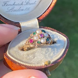May include: A gold ring featuring a cluster of white pearls, small red square-cut stones, and green round-cut stones, set in a band with intricate leaf-like metalwork, presented inside a brown leather jewellery box with the text L. Mumford 1. Fen Court Fenchurch Street London E.C.