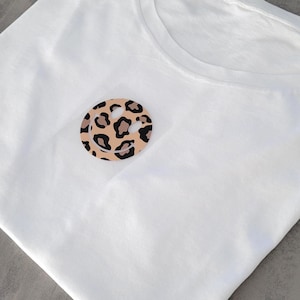 Könnte beinhalten: Weißes T-Shirt mit einem runden Smiley-Gesicht im Leopardenmuster auf der Brust. Das Smiley-Gesicht hat einen beigen Hintergrund mit schwarzen Flecken und einer schwarzen Umrandung. Das Shirt besteht aus weichem, weißem Stoff.