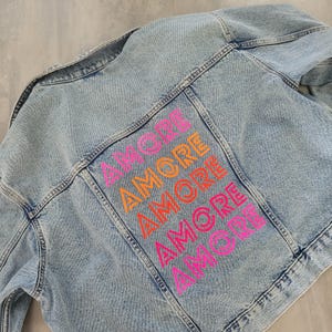 Könnte beinhalten: Hellblaue Jeansjacke mit dem Wort "AMORE" in einem Farbverlauf aus Pink und Orange auf dem Rücken. Die Jacke hat ein klassisches Design mit Kragen, Knopfverschlüssen und Taschen.