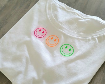 May include: White t-shirt with three smiley face designs in neon pink, orange, and green. The t-shirt is folded and lies on a gray surface. The smiley faces are centered on the shirt.