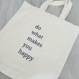 Könnte beinhalten: Beige Canvas-Tragetasche mit dem schwarzen Text "do what makes you happy". Die Tasche hat zwei passende Griffe und besteht aus einem robusten Stoff. Ideal zum Tragen von Büchern, Lebensmitteln oder persönlichen Gegenständen.