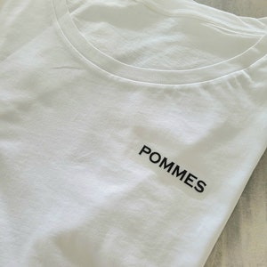 Può includere: Maglietta bianca con un logo nero "POMMES" stampato sul davanti.