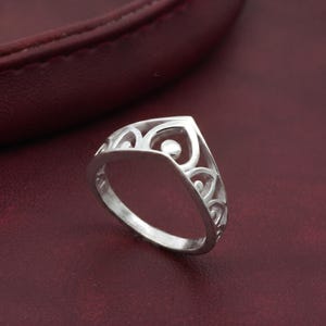 May include: A silver ring with an openwork design. The ring features a heart-shaped top with a small, round accent and decorative swirls. The ring is set against a dark red background.