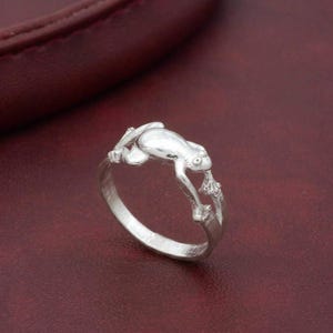 Puede incluir: Un anillo de plata con un diseño detallado de una rana. La rana está esculpida en una pose realista, aferrándose a una rama. El anillo tiene un acabado pulido y los detalles de la rana están bien definidos. El anillo está sobre un fondo rojo oscuro.