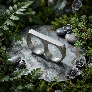 Op de afbeelding: Een zilverkleurige ring voor twee vingers met een rechthoekige balk die twee cirkelvormige banden verbindt. De ring ligt op een getextureerd grijs oppervlak, omgeven door groen en kleine, decoratieve bloemaccenten.