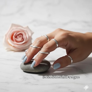 May include: A hand wearing multiple silver rings. The rings are simple bands. The hand is holding a gray stone. A light pink rose is in the background. The nails are painted light blue. The text "BohoBlissfulDesigns" is at the bottom.
