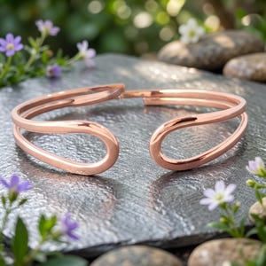 Könnte beinhalten: Roségoldenes Armreif-Armband mit einem doppelten Schlaufendesign. Das offene Armband wird auf einer grauen Oberfläche präsentiert, umgeben von kleinen Steinen und Grün. Die glatte, metallische Oberfläche des Armbands reflektiert das Licht.