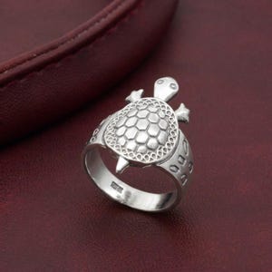 May include: A silver-toned ring featuring a three-dimensional turtle design on the top. The turtle shell has a detailed hexagonal pattern, and the band of the ring is decorated with small, oval-shaped cutouts along the sides of the metal band.