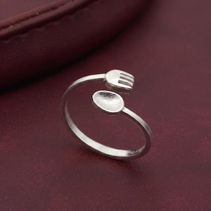 May include: A silver-colored ring featuring a fork and spoon design. The ring has a simple, elegant style, with the fork and spoon forming the top portion of the ring. The ring is set against a dark red background.