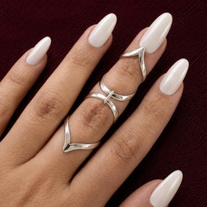 Könnte beinhalten: Ein Set aus drei silberfarbenen Ringen, die an einem einzigen Finger getragen werden. Das Design umfasst einen V-förmigen Ring an der Basis, einen schlichten Ring in der Mitte und einen V-förmigen Gelenkring oben, alle mit glatter Oberfläche.