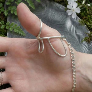 Könnte beinhalten: Ein silberner Daumenring mit einem einzigartigen, offenen Design, der durch eine zarte Kette mit einem Armband verbunden ist. Der Ring hat eine geschwungene, abstrakte Form, die den Daumen umschließt. Der Schmuck wird vor einem natürlichen, moosigen Hintergrund präsentiert.