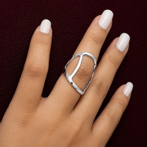 May include: A polished silver-tone ring featuring an open, asymmetrical geometric design. The band consists of a smooth, curved metal frame that wraps around the finger, creating a large, hollow center space with a vertical bar dividing the shape.
