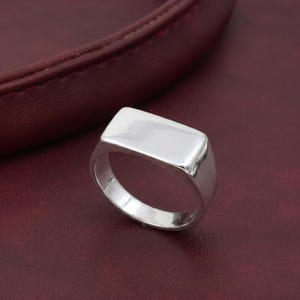 Op de afbeelding: Een glanzende zilveren zegelring met een rechthoekige voorkant. De ring is geplaatst tegen een donkerrode achtergrond met leertextuur. Het oppervlak van de ring reflecteert licht, wat de gepolijste afwerking benadrukt.