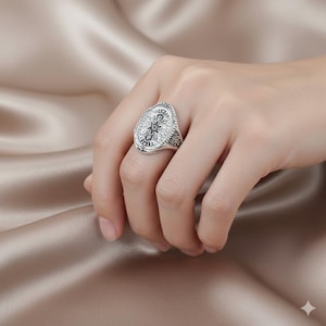 Könnte beinhalten: Ein silberfarbener Ring mit ovaler Oberfläche, der ein detailliertes Blumenmuster zeigt. Der Ring hat ein strukturiertes Band und wird am Finger getragen. Der Hintergrund ist ein weicher, neutraler Stoff.