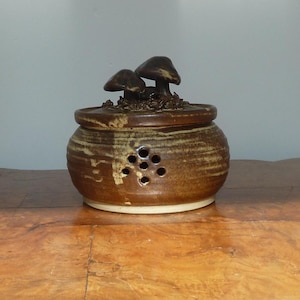 May include: A brown ceramic jar with a lid decorated with two mushroom figures. The jar is round with several small holes and a light base. The surface has a textured, earthy appearance, reminiscent of a woodland theme.