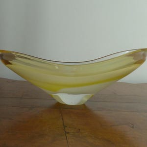May include: A clear glass bowl with a yellow and white swirl design. The bowl has a wide, curved shape and sits on a clear, faceted base. The bowl is displayed on a wooden surface.