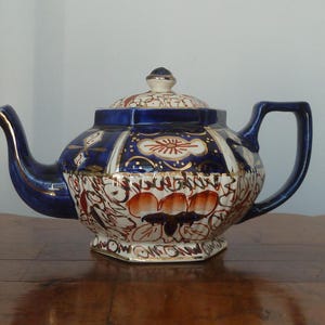 May include: A blue and white teapot with gold accents and floral designs. The teapot has a round body and a spout on the side. The handle is on the opposite side of the spout.