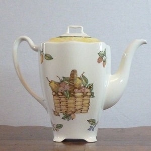 May include: A white ceramic teapot with a yellow rim and a floral design featuring a basket of fruit. The teapot has a handle and a spout.