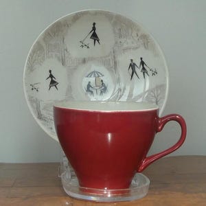 May include: A red teacup and saucer set. The teacup is a deep red colour, and the saucer is white with a black illustration of people walking dogs and dining at a cafe. The teacup is sitting on a clear plastic coaster.