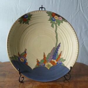 May include: A large, cream-coloured ceramic bowl with a blue rim and a floral design. The bowl features a stylised floral pattern with orange, yellow, blue, and green accents. The bowl is on a wooden surface.
