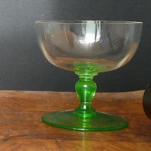 May include: A clear glass champagne coupe with a bright green stem and base. The bowl is wide and shallow, with a bulbous stem design. The coupe sits on a round base. The glass is on a wooden surface.