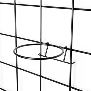 Pode incluir: Suporte para plantas de metal preto com um anel circular para segurar um vaso. O suporte é fixado a uma grelha de metal preto.