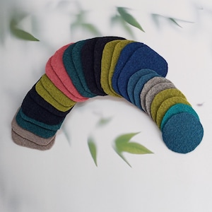 Könnte beinhalten: Eine Sammlung farbenfroher Filzuntersetzer in verschiedenen Formen und Farbtönen, darunter Rosa, Grün, Blau und Grau. Die Untersetzer sind in einer gebogenen Form angeordnet, was auf eine dekorative oder organisatorische Präsentation hindeutet. Der Hintergrund ist ein weiches Weiß mit verschwommenen grünen Blättern.