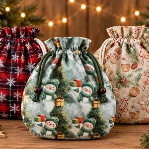 Könnte beinhalten: Drei Stoffbeutel mit Kordelzug und weihnachtlichen Motiven. Der linke Beutel ist rot-schwarz kariert mit weißen Schneeflocken. Der mittlere Beutel ist hellgrün mit Schneemännern und Bäumen. Der rechte Beutel ist beige mit Lebkuchen.