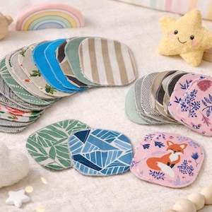 Könnte beinhalten: Eine Sammlung wiederverwendbarer Babytücher mit verschiedenen Mustern, darunter Blumen-, geometrische und gestreifte Designs. Die Tücher sind zusammen mit anderen Spielzeugen für Babys angeordnet, wie einem sternförmigen Plüschtier, Holzbausteinen und einem wolkenförmigen Plüschtier.