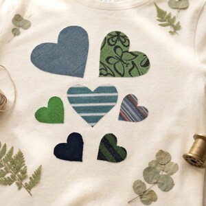 Könnte beinhalten: Ein cremefarbenes T-Shirt mit sieben herzförmigen Stoffaufnähern in verschiedenen Mustern und Texturen, darunter einfarbiges Blau, Grün und Marineblau sowie gestreifte und mit Schmetterlingen bedruckte Designs auf der Vorderseite.