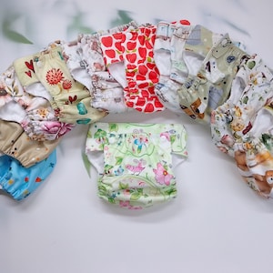 Könnte beinhalten: Eine Sammlung bunter Stoffwindeln mit verschiedenen Mustern, darunter Blumen-, Tier- und geometrische Designs. Die Windeln sind in einem Halbkreis auf weißem Hintergrund angeordnet.