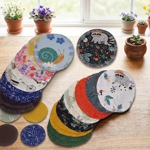 Könnte beinhalten: Eine Sammlung wiederverwendbarer, runder Wattepads in verschiedenen Farben und Mustern, darunter Blumen-, Schnecken- und Waschbären-Designs. Die Pads sind auf einem Holztisch angeordnet, zusammen mit Topfpflanzen.