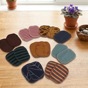 Könnte beinhalten: Eine Ansammlung farbenfroher, quadratischer Stoffuntersetzer, die auf einer Holzoberfläche angeordnet sind. Die Untersetzer weisen verschiedene Muster auf, darunter Streifen, florale Designs und unifarbene Farben. Eine kleine Topfpflanze und eine Holzschale mit Steinen sind ebenfalls zu sehen.