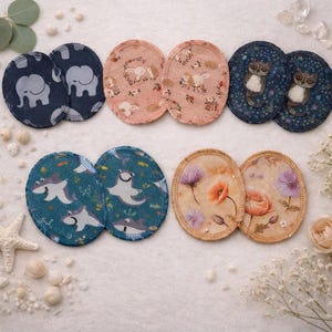 Könnte beinhalten: Acht kreisförmige Stoffpads, angeordnet in zwei Reihen. Die obere Reihe zeigt zwei dunkelblaue Pads mit Elefantenmuster, zwei hellrosa Pads mit Hasenmuster und zwei dunkelblaue Pads mit Eulenmuster. Die untere Reihe enthält zwei türkisfarbene Pads mit Hai-Motiv und zwei beige Pads mit Blumenmuster.