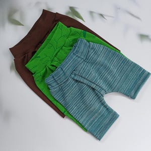 Pu&ograve; includere: Tre paia di pantaloncini per bambini in marrone, verde e blu. Il paio marrone &egrave; tinta unita, il paio verde ha un motivo a foglie e il paio blu ha un motivo a righe. I pantaloncini sono impilati su una superficie bianca.
