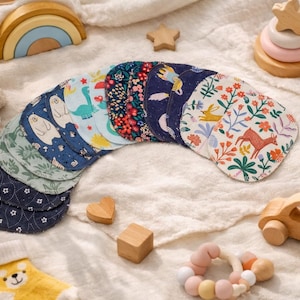 Könnte beinhalten: Eine Sammlung kreisförmiger Stoffpads, die fächerförmig angeordnet sind und verschiedene Muster wie Blumenprints, Waldtiere, geometrische Formen und dunkelblaue Hintergründe mit weißen Sternen auf einer weichen, strukturierten Stoffoberfläche zeigen.